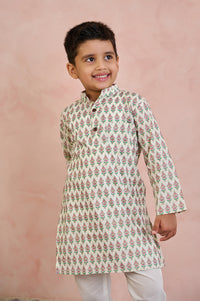 A young boy wearing a cream-colored kurta with a green and pink leaf pattern, paired with white trousers, standing against a textured pink background.