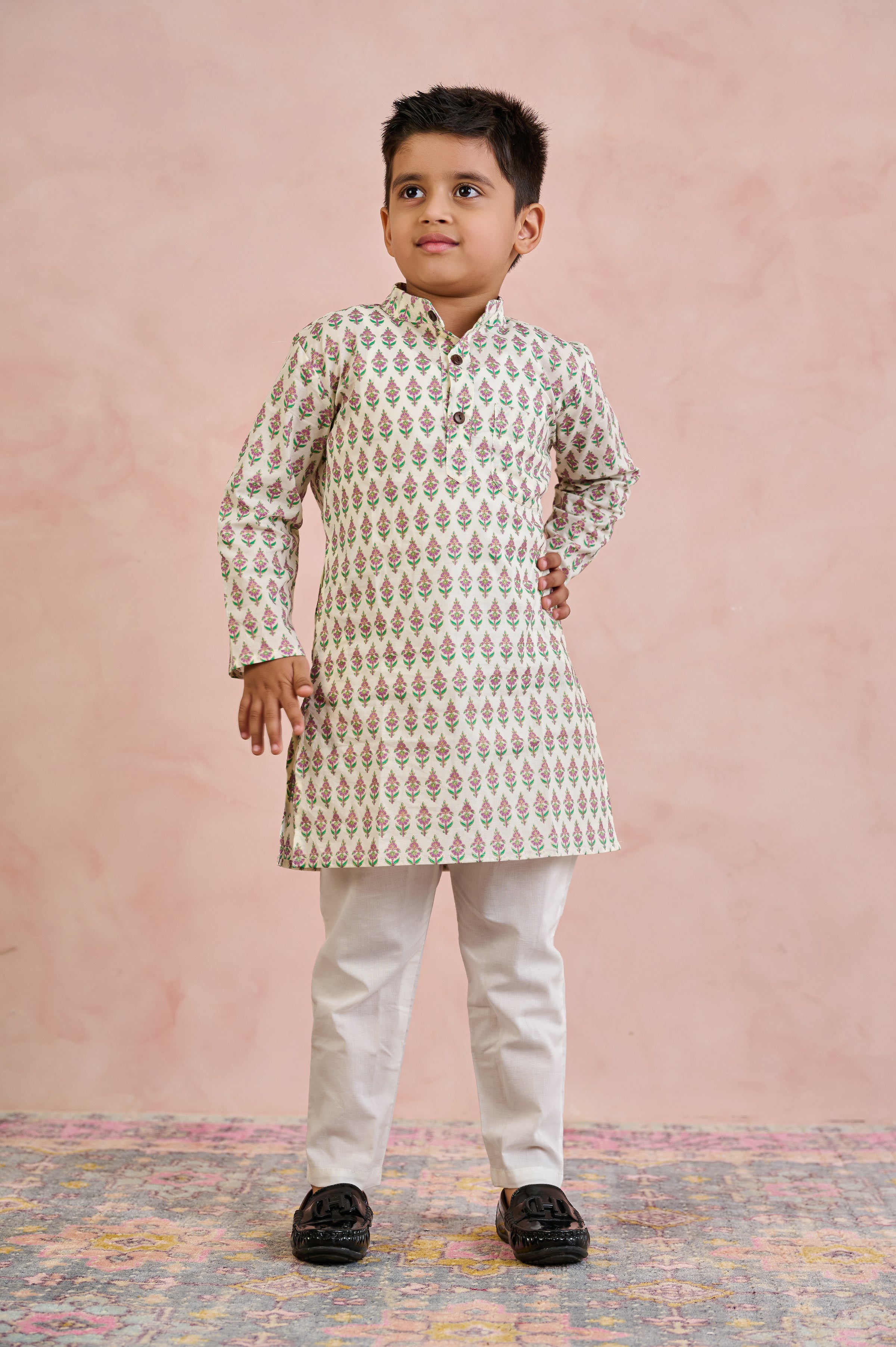 A young boy modeling a traditional Indian outfit consisting of a cream-colored kurta with a green and gold geometric pattern, paired with off-white trousers and black shoes, standing against a textured pink backdrop.