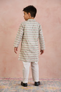 A young boy viewed from the back wearing a cream-colored kurta with a repeating green and pink floral block print, paired with off-white trousers and black shoes, standing against a textured pink wall.