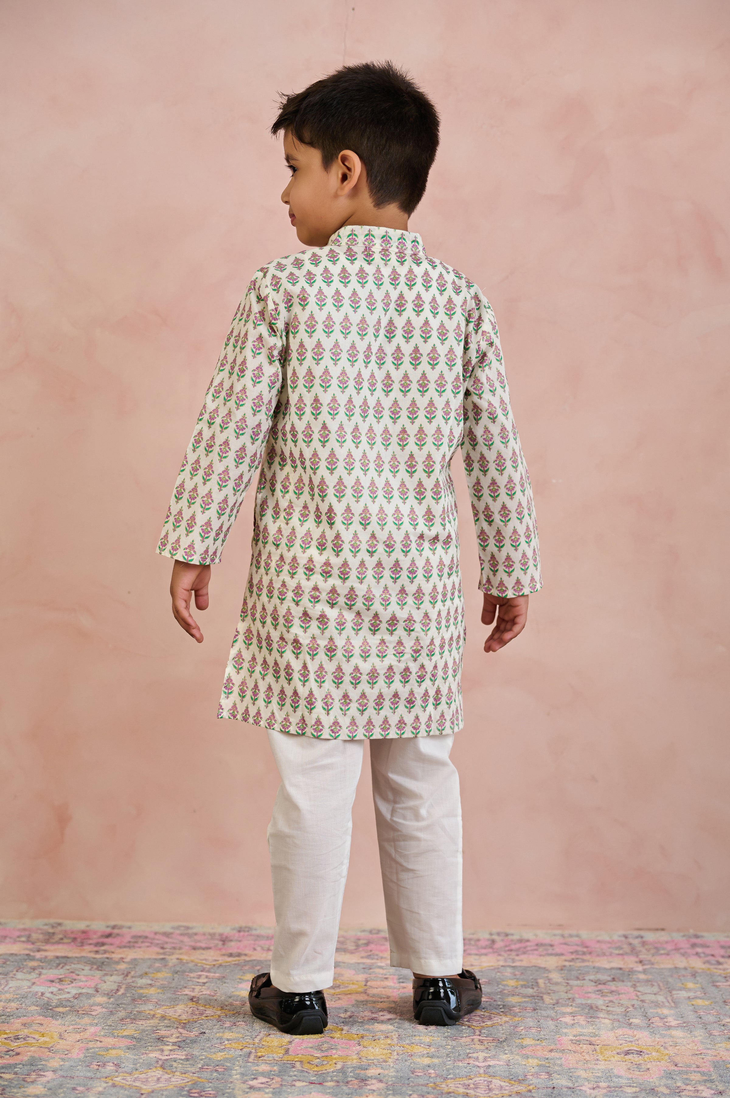 A young boy viewed from the back wearing a cream-colored kurta with a repeating green and pink floral block print, paired with off-white trousers and black shoes, standing against a textured pink wall.