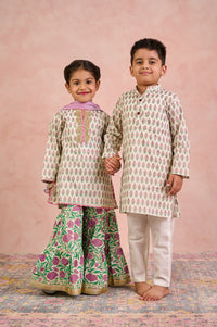 Two children modeling traditional Indian ethnic wear against a textured pink backdrop. The girl wears a beige kurta with a pink collar and green floral sharara pants, while the boy wears a matching beige kurta with white pants.