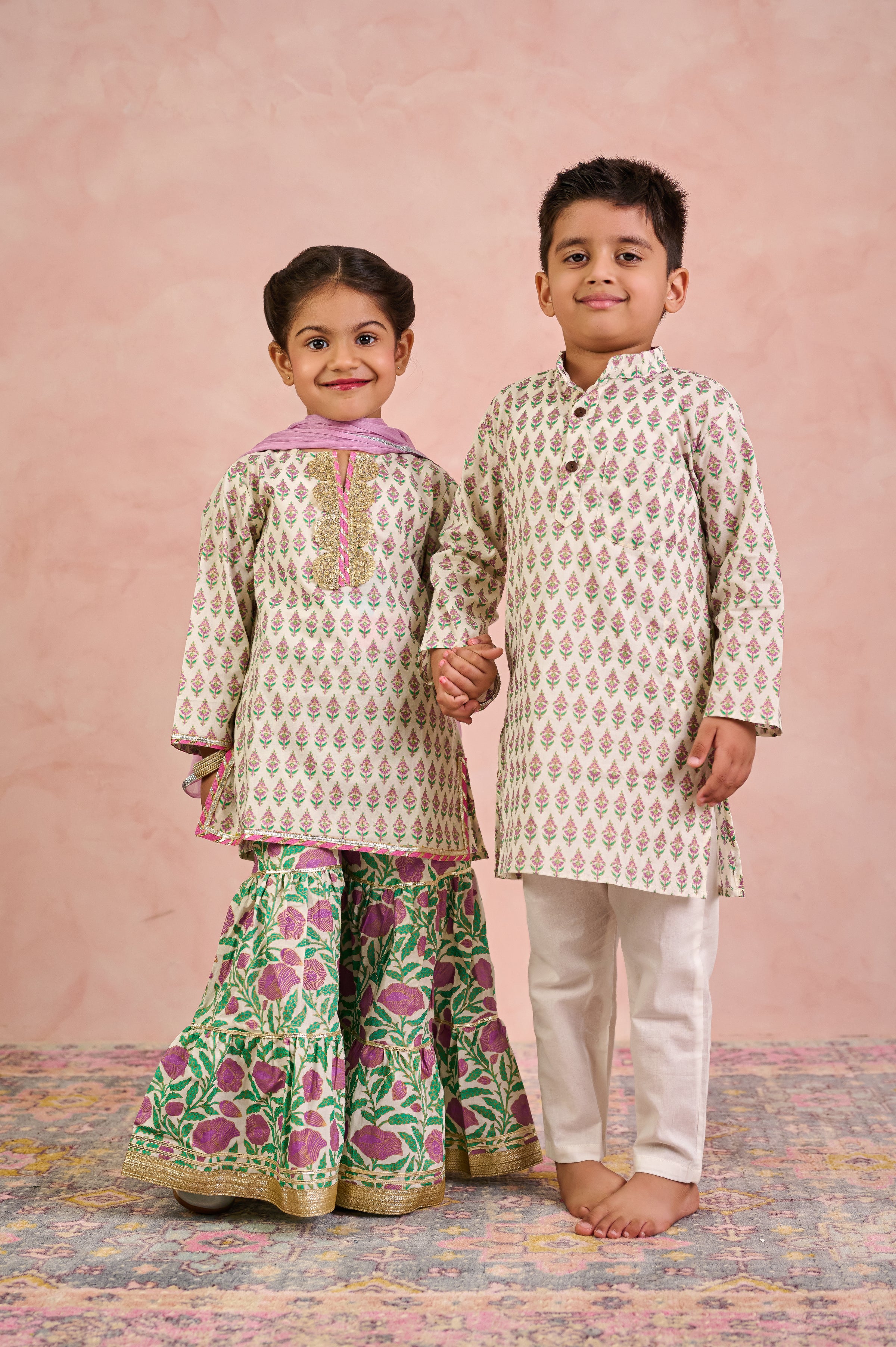 Two children modeling traditional Indian ethnic wear against a textured pink backdrop. The girl wears a beige kurta with a pink collar and green floral sharara pants, while the boy wears a matching beige kurta with white pants.