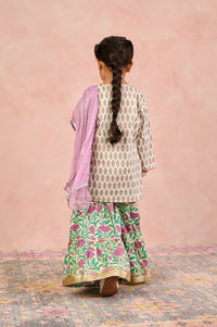 A young girl viewed from behind wearing a traditional Indian outfit with a cream kurta featuring a green and pink floral print, matching green and pink flared pants, and a solid light pink dupatta, set against a soft pink textured wall.