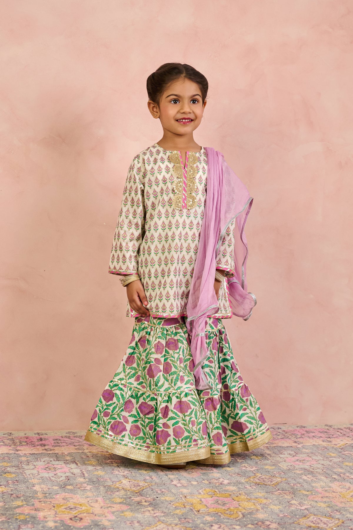 A young girl modeling a traditional Indian outfit consisting of a cream kurta with green and pink floral prints, a matching green flared skirt with pink floral prints and gold borders, and a light pink dupatta, set against a textured pink backdrop.