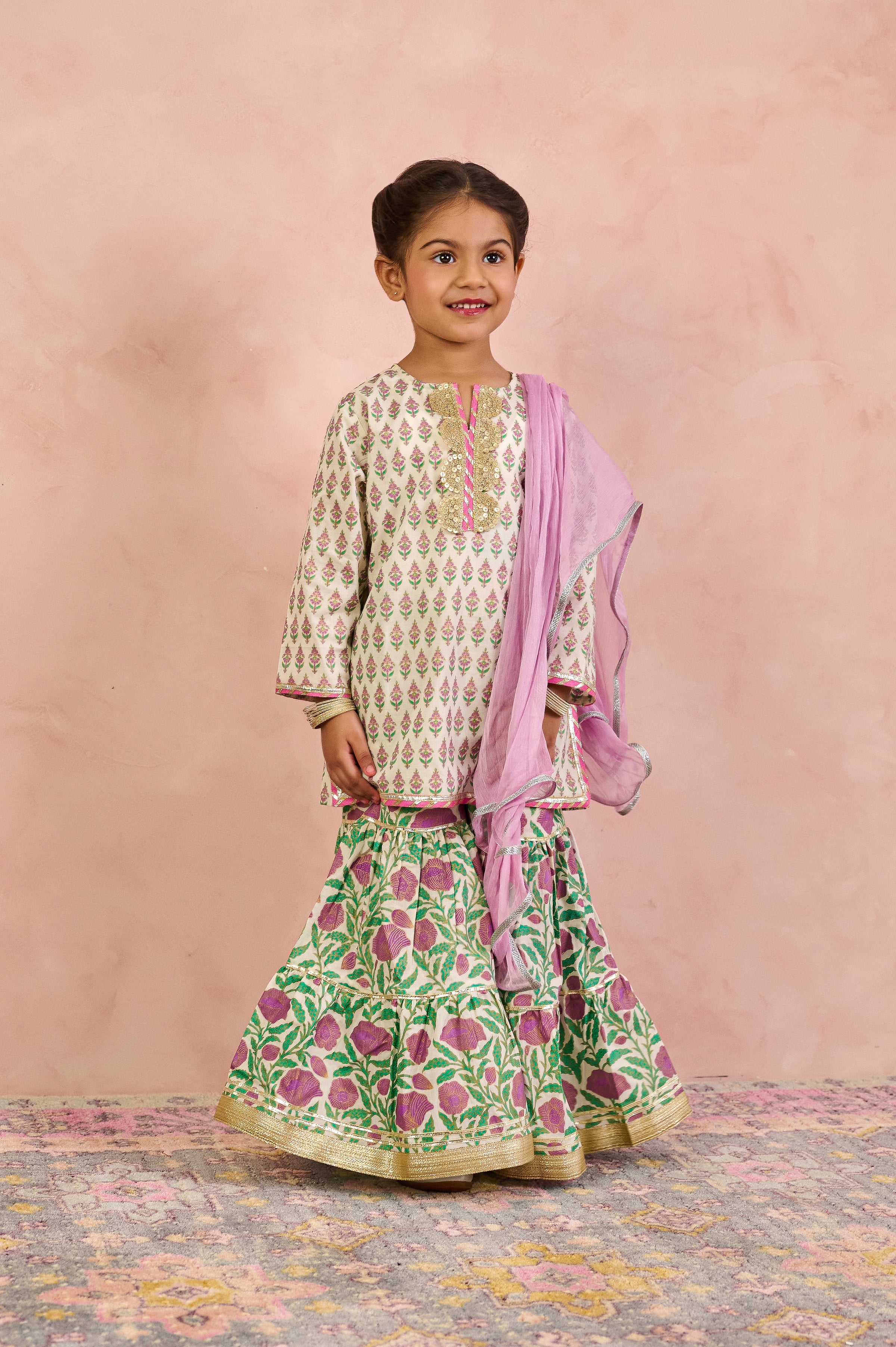 A young girl modeling a traditional Indian outfit consisting of a cream kurta with green and pink floral prints, a matching green flared skirt with pink floral prints and gold borders, and a light pink dupatta, set against a textured pink backdrop.