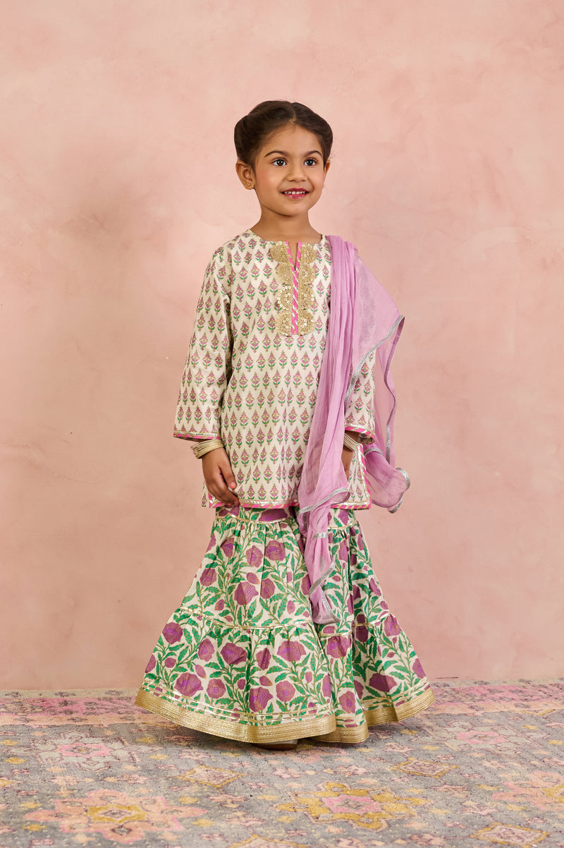 A young girl modeling a traditional Indian outfit consisting of a cream kurta with green and pink floral prints, a matching green flared skirt with pink floral prints and gold borders, and a light pink dupatta, set against a textured pink backdrop.