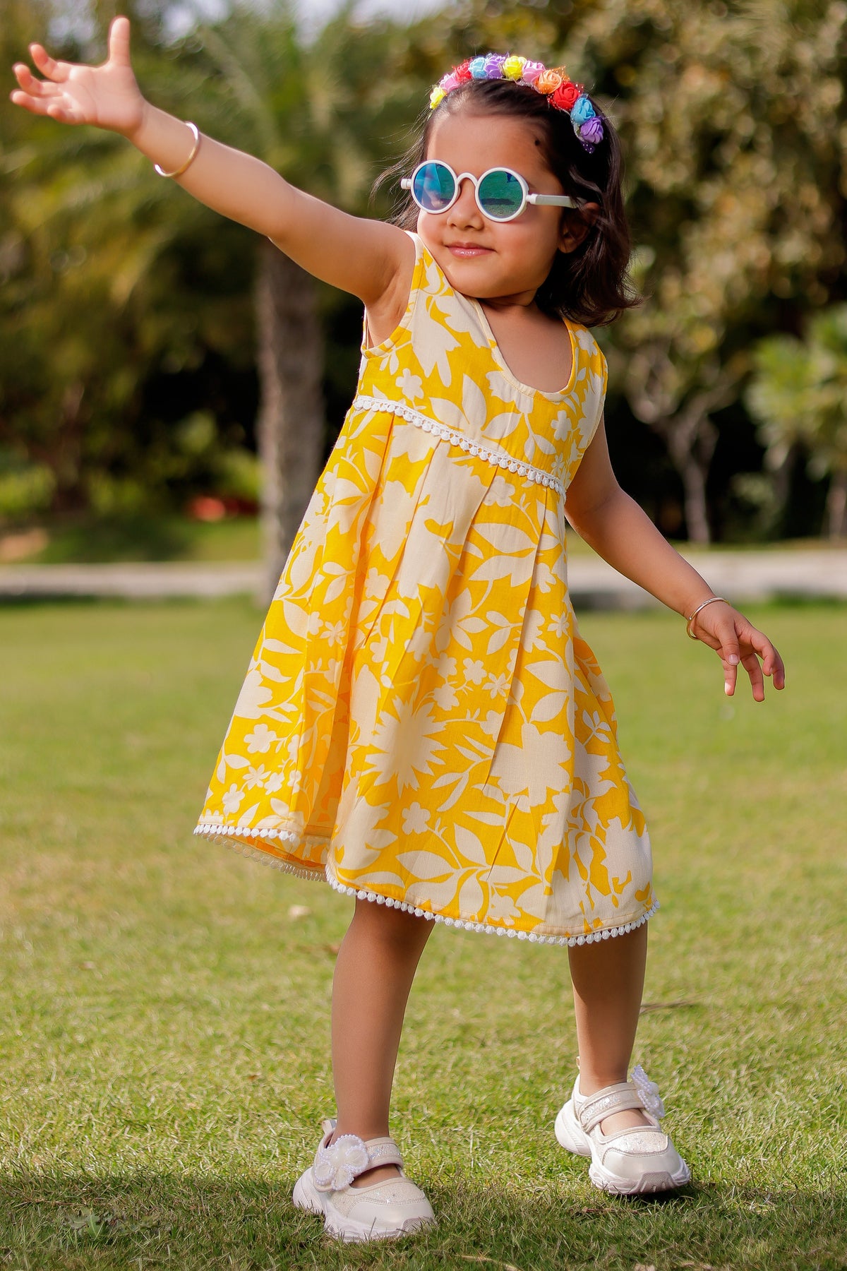 Child in a yellow dress with floral pattern standing on grass with trees in the background