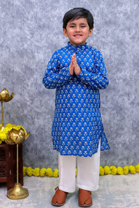 A young boy wearing a royal blue kurta with a white allover floral pattern, paired with white pants and brown sandals, standing in a prayer pose against a textured gray backdrop.
