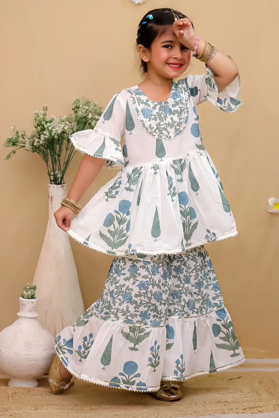 A young girl models a two-piece traditional outfit featuring a white peplum top and matching flared skirt with a blue and green botanical pattern, set against a warm beige background with decorative vases.