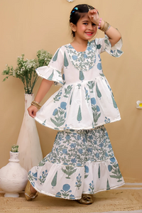 A young girl models a two-piece traditional outfit featuring a white peplum top and matching flared skirt with a blue and green botanical pattern, set against a warm beige background with decorative vases.