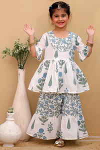 A young girl modeling a white two-piece traditional outfit with blue and green floral block prints, standing in front of a beige wall with decorative vases.