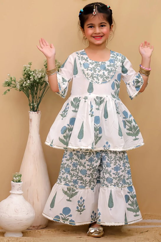 A young girl modeling a white two-piece traditional outfit with blue and green floral block prints, standing in front of a beige wall with decorative vases.