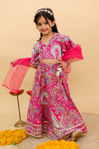 A young girl modeling a vibrant pink and multicolor paisley printed traditional Indian outfit, consisting of a crop top, flared skirt, and matching dupatta, set against a solid beige backdrop.