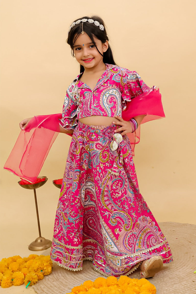 A young girl modeling a vibrant pink and multicolor paisley printed traditional Indian outfit, consisting of a crop top, flared skirt, and matching dupatta, set against a solid beige backdrop.