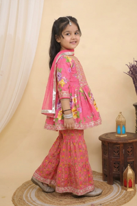 A young girl modeling a  Pink Embroidered  Floral Peplum Style Kurta with Sharara and Dupatta from Desi Trove, standing on a round jute rug in a warm beige studio setting.