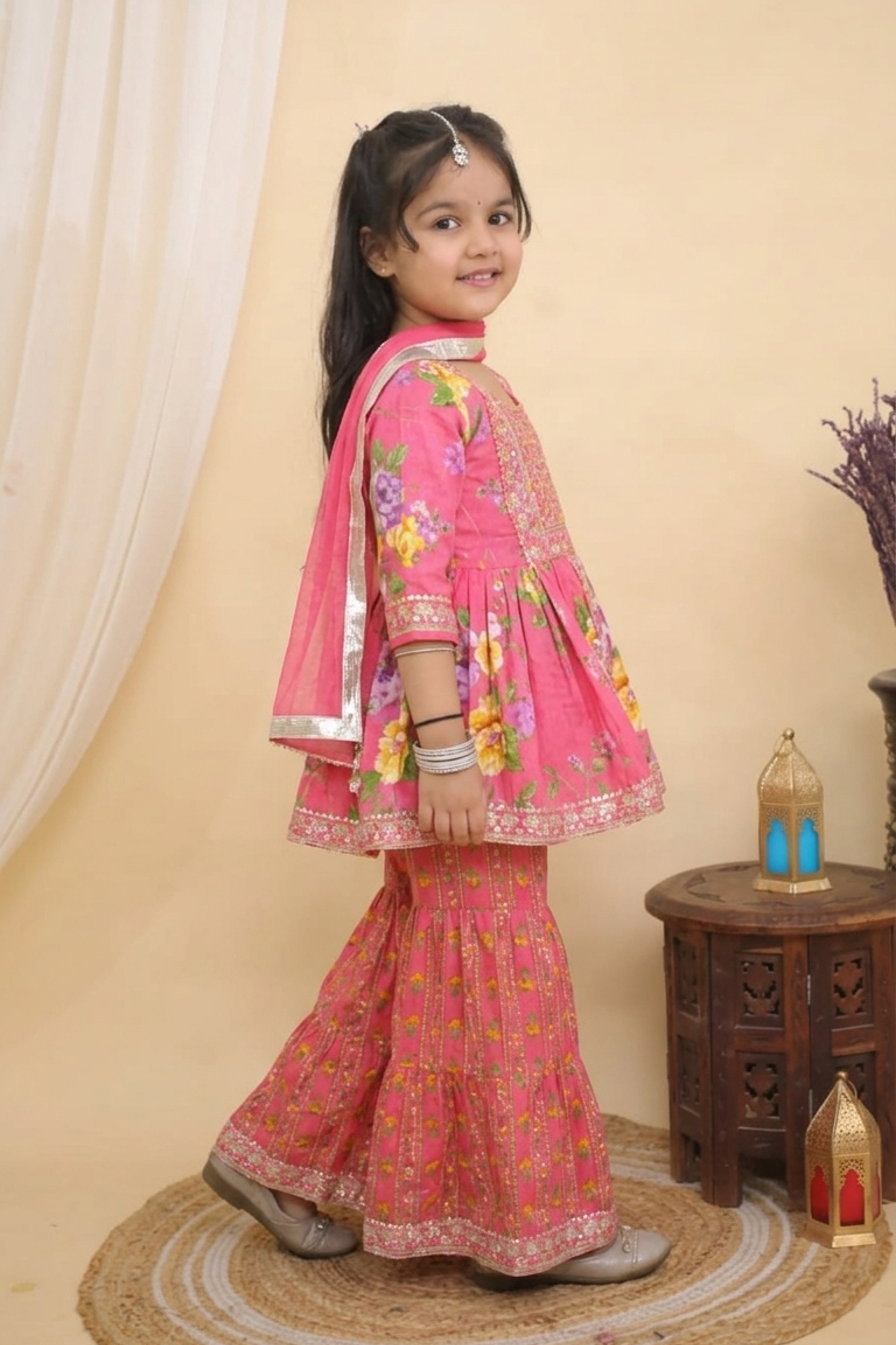 A young girl modeling a  Pink Embroidered  Floral Peplum Style Kurta with Sharara and Dupatta from Desi Trove, standing on a round jute rug in a warm beige studio setting.