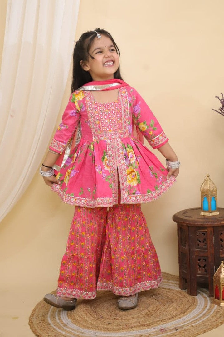 A young girl modeling a Pink Embroidered  Floral Peplum Style Kurta with Sharara and Dupatta from Desi Trove, standing on a woven mat in a decorated indoor setting.