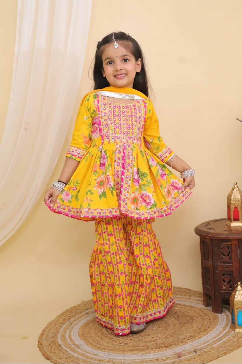 Young girl in a Yellow Embroidered Floral Peplum Style Kurta with Sharara and Dupatta from Desi Trove standing in a room with decorative elements.