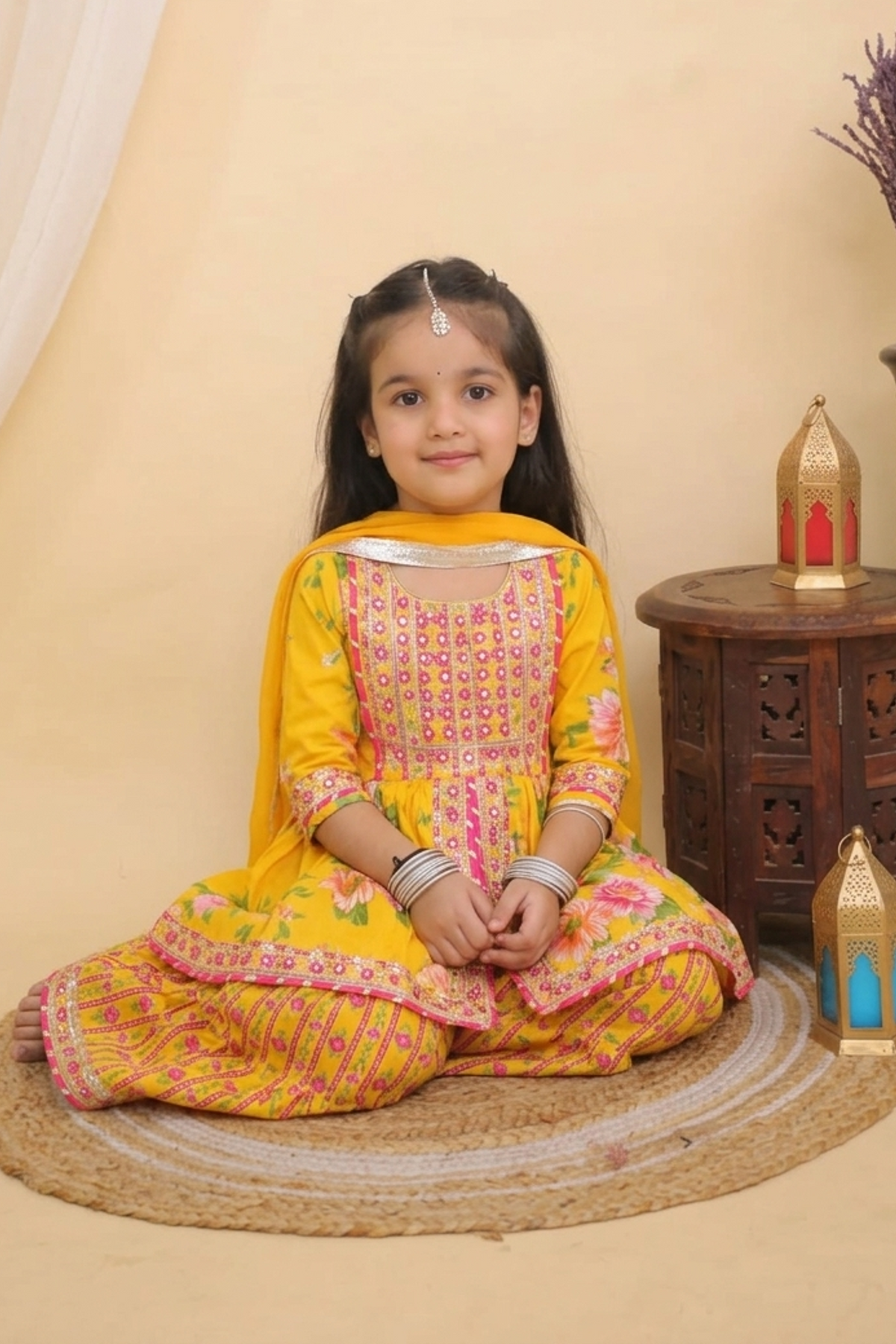 A young girl sits cross-legged on a round jute mat wearing a Yellow Embroidered Floral Peplum Style Kurta with Sharara and Dupatta
 from Desi Trove. She is posed in a studio setting with a beige backdrop, a small wooden side table, decorative lanterns, and a vase with dried flowers.