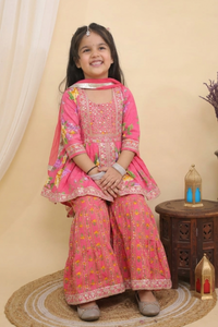 A young girl smiling while wearing a Pink Embroidered  Floral Peplum Style Kurta with Sharara and Dupatta from Desi Trove , seated on a round woven mat in a studio setting with a small wooden side table and decorative lanterns.