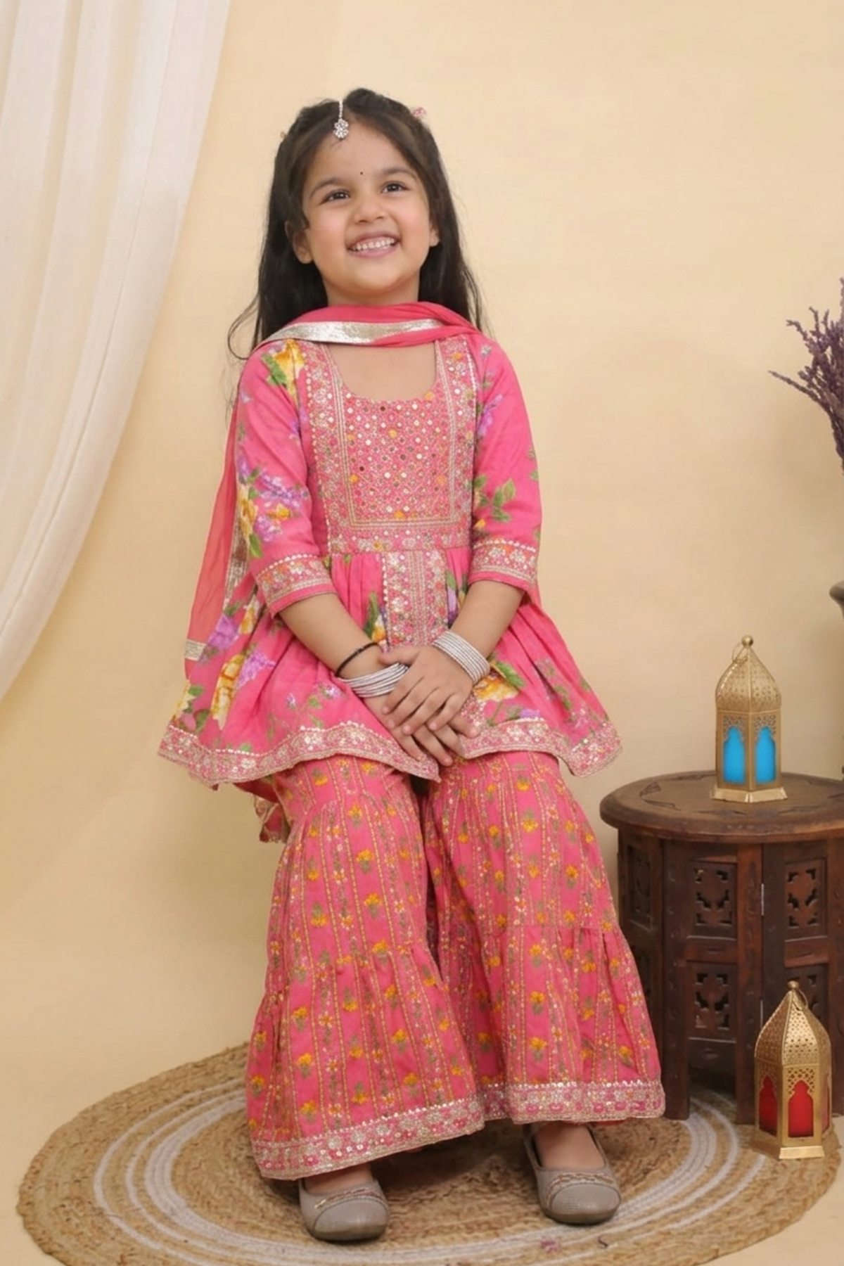 A young girl smiling while wearing a Pink Embroidered  Floral Peplum Style Kurta with Sharara and Dupatta from Desi Trove , seated on a round woven mat in a studio setting with a small wooden side table and decorative lanterns.