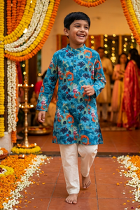 A young boy smiling and walking barefoot down a decorated aisle, wearing a vibrant blue patterned kurta with white pants. The background features colorful marigold and white flower garlands and warm string lights.