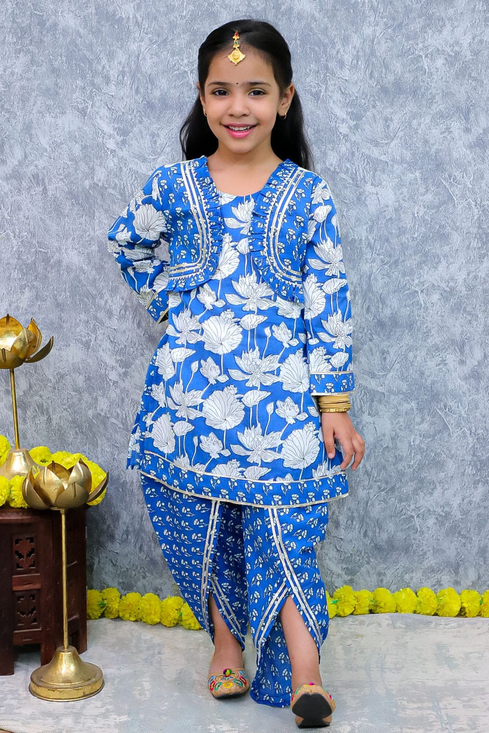 A young girl modeling a blue and white printed traditional Indian outfit with a kurta and matching pants, standing against a textured gray backdrop with decorative props.