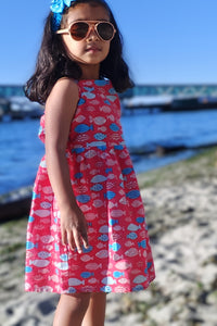 Young girl in a red dress with fish pattern standing by a waterfront.