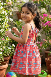 Young girl in a red fish print dress standing in a garden with flowers and plants.
