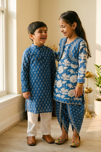A young boy and girl wearing matching blue and white traditional Indian outfits, standing together in a brightly lit room with a window and wooden floor.