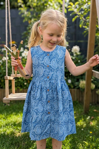 Young girl in a blue paisley print cotton  dress standing next to a wooden swing with a toy figure, outdoors.