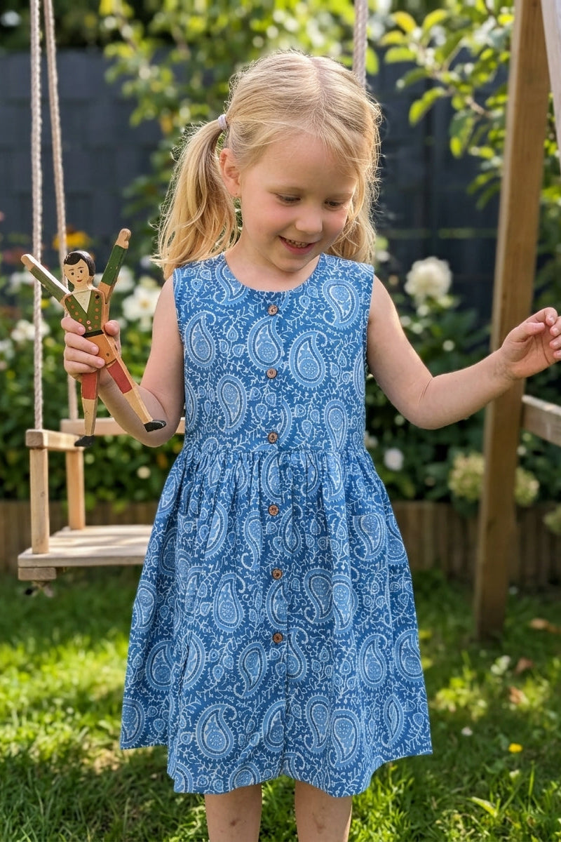 Young girl in a blue paisley print cotton  dress standing next to a wooden swing with a toy figure, outdoors.