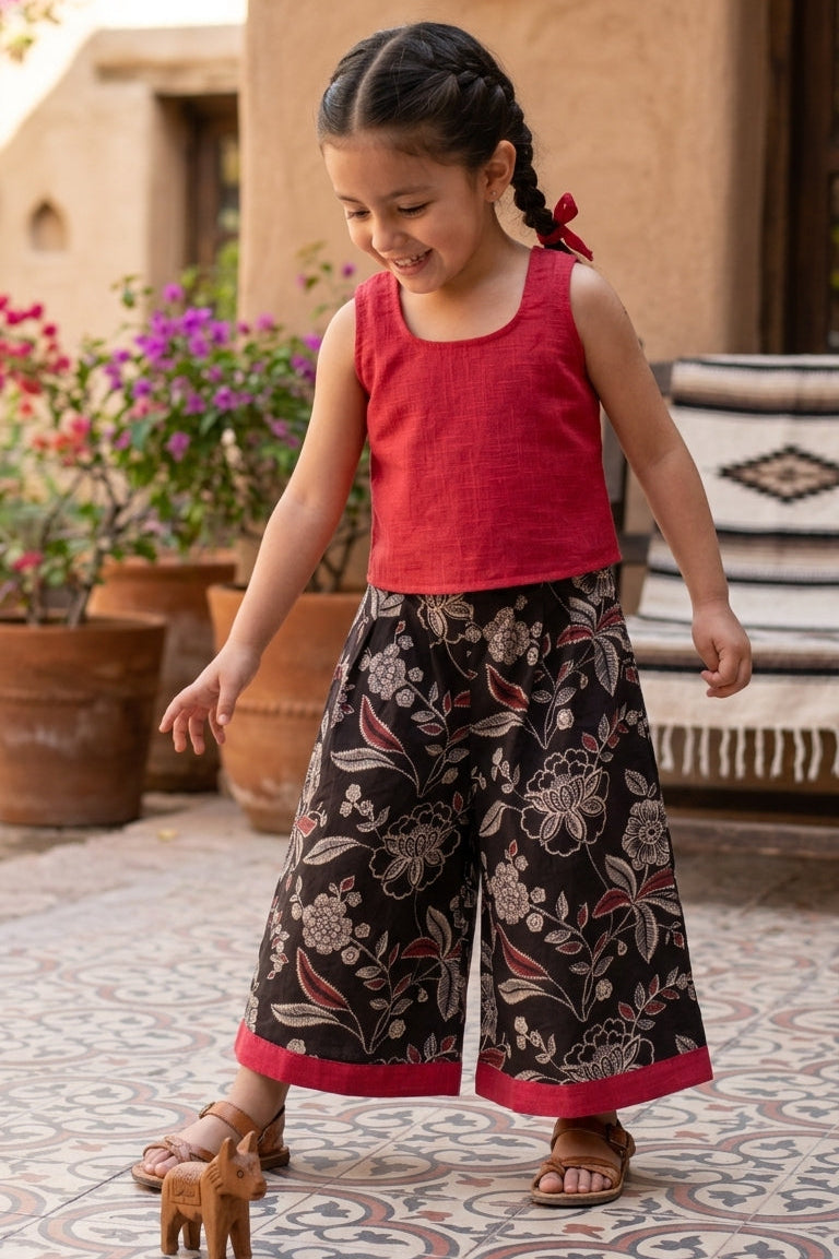 Young girl in a red top and patterned culotte standing on a patio with plants and furniture in the background.