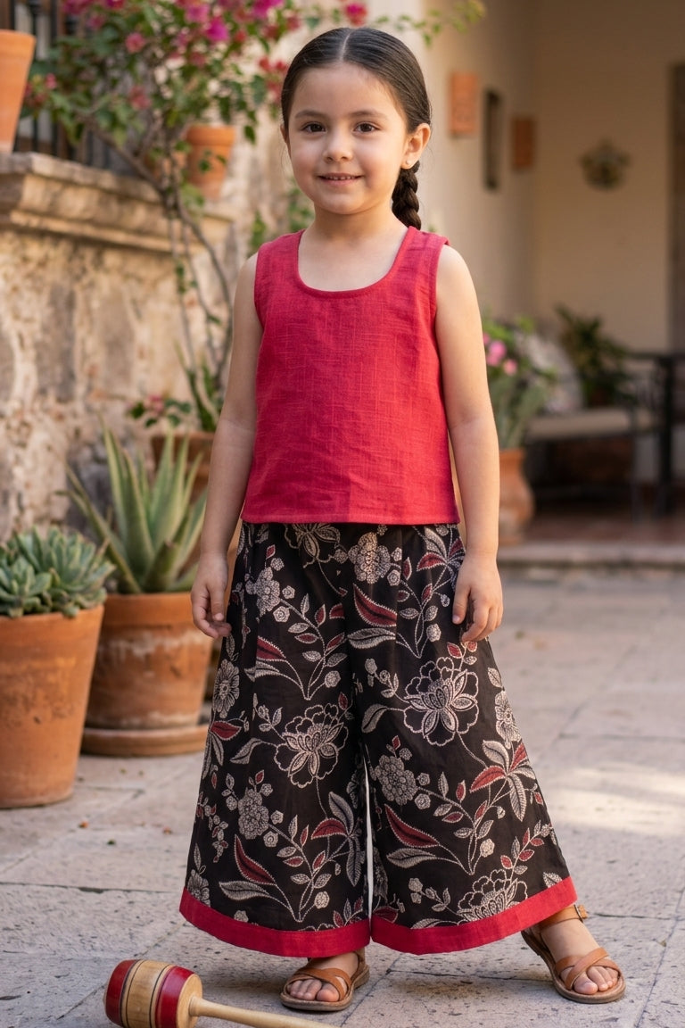 Young girl in a red top and patterned culotte standing outdoors with potted plants and flowers in the background.