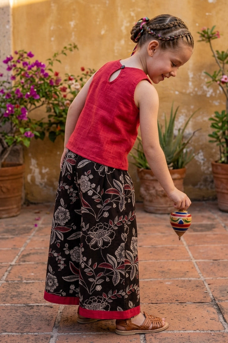 Young girl in a red top and black floral culotte standing on a tiled floor with plants in the background.