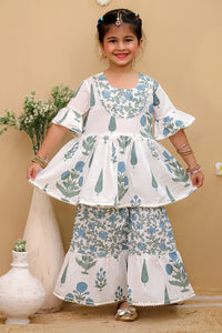 A young girl modeling a white two-piece traditional outfit with blue and green floral prints, standing against a beige wall with decorative vases.